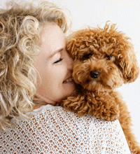 Kvinde står med lille brun hund over skulderen, og læner sit smilende ansigt mod hunden.