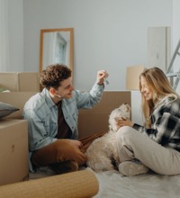 Ungt par sidder på gulvet mellem flyttekasser, med deres hund og et sæt nøgler.