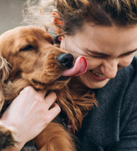 Kvinde står med en brun hund i armene, mens den er i færd med at slikke hende i hovedet.