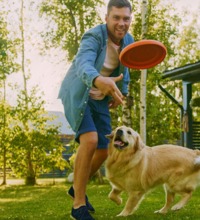 Mand og hund i haven, hvor manden kaster en frisbee til hunden.